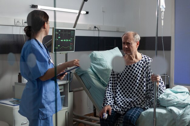 Personal médico con estetoscopio interrogando a un hombre mayor enfermo sentado en la cama con goteo intravenoso, con expresión dolorosa