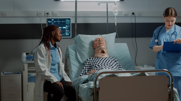 Personal médico multiétnico que controla la atención médica del paciente en la cama de la sala de hospital en cuidados intensivos. Médico afroamericano ayudando a anciano con collarín cervical y tubo de oxígeno