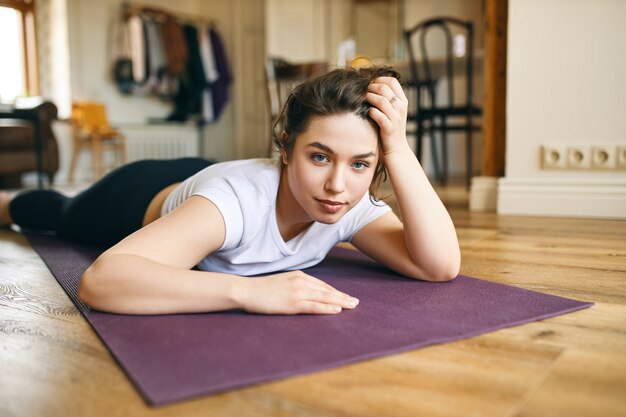 Personas, actividad física, deportes, fitness y distanciamiento social. Mujer deportiva joven cansada acostada boca abajo, relajándose después de un entrenamiento intensivo en casa, mirando a la cámara, habiendo agotado la mirada