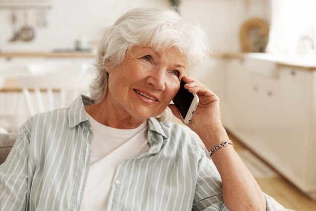 Personas, aparatos electrónicos modernos, tecnología y comunicación. Mujer mayor de edad con pelo gris corto disfrutando de una agradable conversación telefónica, sentada en el sofá, sosteniendo el móvil en su oído y sonriendo