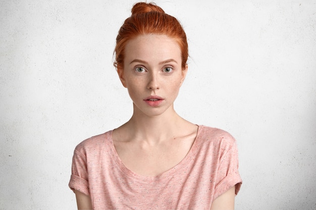 Foto gratuita personas, concepto de belleza natural. grave atractiva mujer joven de ojos de avaricia con cabello pelirrojo, vestida con una camiseta rosa casual, centrada en algo en el frente