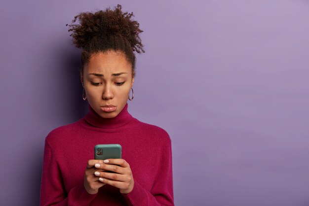 Personas, concepto de emociones infelices. Triste mujer decepcionada recibió malos sms, sostiene el teléfono inteligente y se concentra en la pantalla con insatisfacción, usa poloneck burdeos, posa en interiores, se siente sola