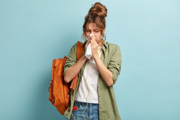 Personas, concepto de enfermedad. Enferma mujer de cabello oscuro estornuda en pañuelo, lleva mochila