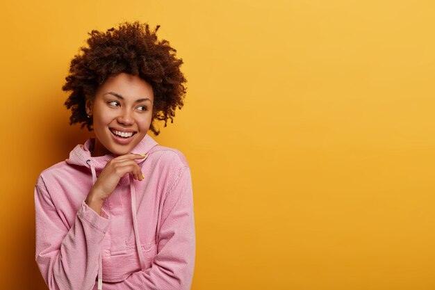 Personas, concepto de estilo de vida. Mujer de piel oscura positiva feliz de encontrar buenas oportunidades para el trabajo futuro, sostiene la barbilla mira hacia otro lado con una amplia sonrisa escucha noticias maravillosas se siente optimista, posa en una pared amarilla