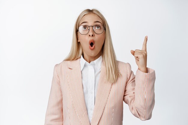 Personas corporativas Mujer de negocios sorprendida señalando y mirando hacia arriba con expresión de cara impresionada que muestra el fondo blanco del anuncio