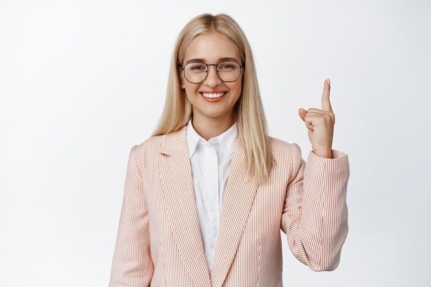 Personas corporativas Vendedora profesional en traje y anteojos sonriendo señalando con el dedo hacia arriba mostrando el fondo blanco del anuncio