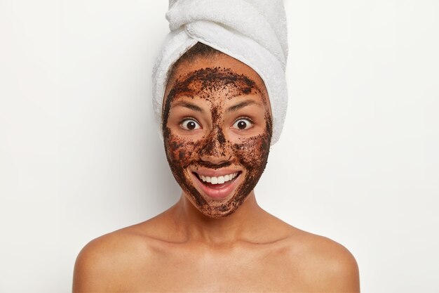 Personas, cuidado de la piel y concepto de belleza. Sonriente modelo femenino de piel oscura limpia la piel con un exfoliante de café, se ve con alegría, sonríe ampliamente, ha envuelto una toalla en la cabeza