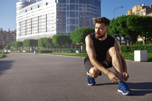 Personas, deportes, estilo de vida activo y concepto de fitness. Retrato de joven deportista cansado con peinado elegante y barba espesa que descansa durante el ejercicio cardiovascular, sentado sobre hormigón y atando cordones de zapatos