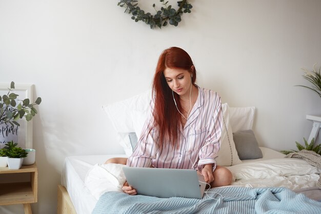 Personas, descanso, relajación y concepto de tecnología moderna. Imagen de hermosa mujer pelirroja en pijama de rayas sentada en la cama deshecha usando una computadora portátil y auriculares mientras ve una película en línea