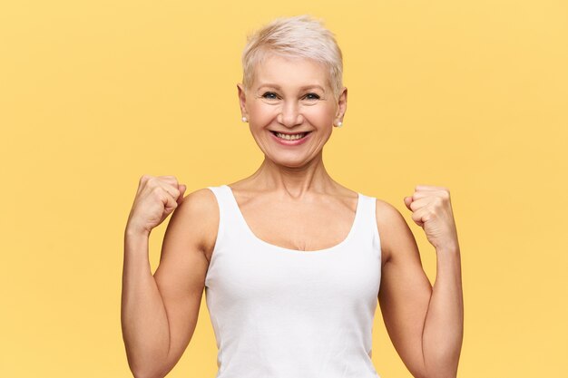 Personas, edad, bienestar y concepto de salud. Atractiva y elegante mujer madura con camiseta blanca sin mangas mostrando sus brazos musculosos, apretando los puños y sonriendo ampliamente, con una mirada feliz y enérgica