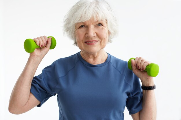 Personas, edad, deportes y concepto de estilo de vida activo. Imagen de mujer jubilada madura positiva feliz en camiseta haciendo ejercicio con pesas en el gimnasio. Entrenamiento femenino senior emocionado con pesas