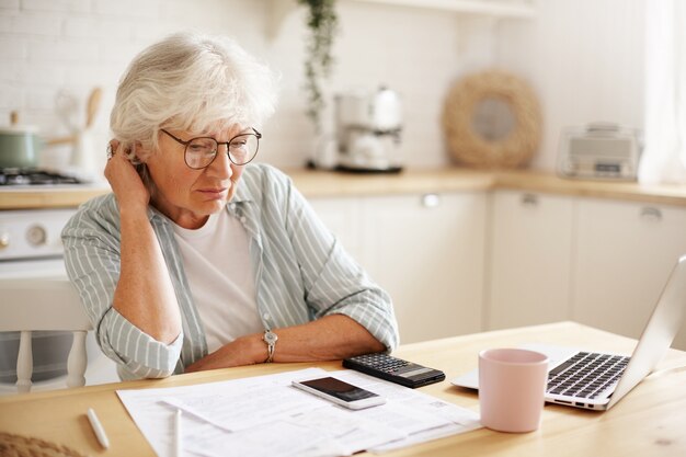 Personas, edad, tecnología y finanzas. Mujer jubilada infeliz deprimida pagando facturas domésticas en línea, esforzándose por llegar a ambos extremos, sentada en la mesa de la cocina, rodeada de papeles, usando aparatos