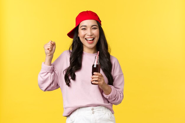 Personas emociones, bebidas y concepto de ocio de verano. Chica asiática feliz entusiasta disfrutando de su refresco, bebiendo bebidas y bailando, sonriendo alegre, de pie fondo amarillo