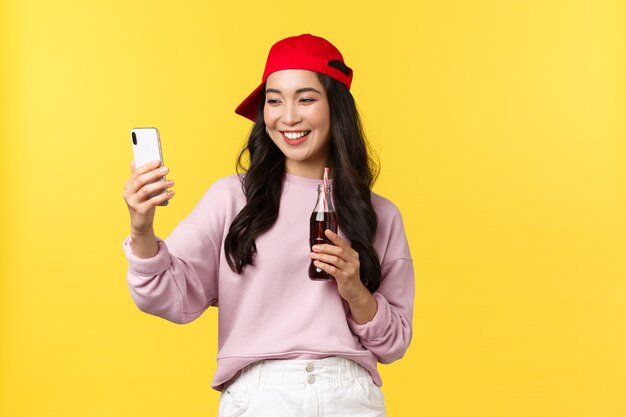 Personas emociones, bebidas y concepto de ocio de verano. Elegante y linda bloguera asiática en gorra roja, tomando selfie con teléfono inteligente, bebiendo refrescos y fotografiándose a sí misma.