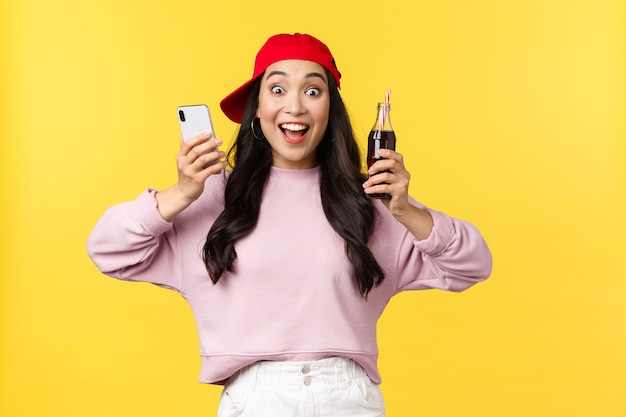 Personas emociones, bebidas y concepto de ocio de verano. Emocionada y entusiasta bastante adolescente asiática que se regocija con el nuevo sabor de la soda, recomienda beber, sostener el teléfono móvil y la botella