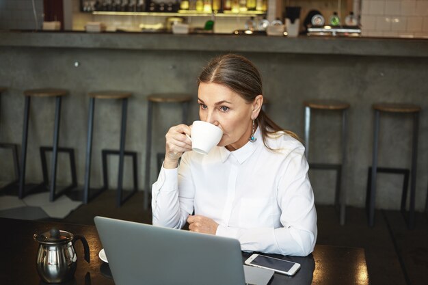 Personas, estilo de vida moderno, tecnologías, concepto de comunicación y ocio. Señora jubilada reflexiva seria con el pelo gris que usa la PC portátil para el trabajo remoto mientras toma un café en la cafetería sola