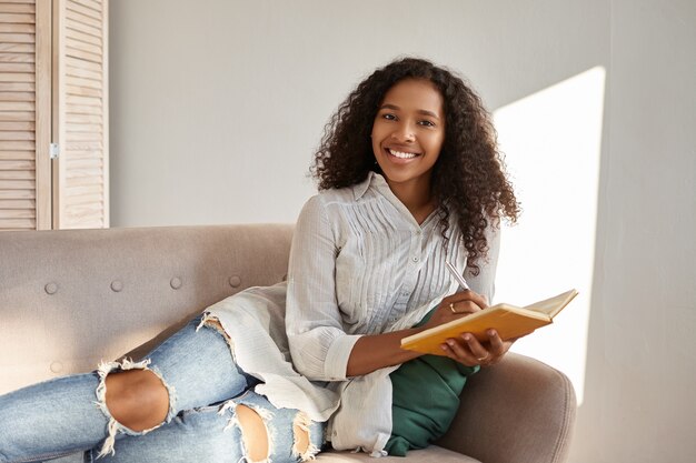 Personas, estilo de vida, ocio, afición y descanso. Adorable encantadora joven mujer de piel oscura con peinado afro relajándose en un cómodo sofá gris sonriendo, anotando metas y planes en el diario