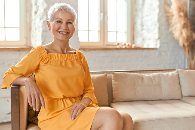 Personas, estilo de vida, ocio, jubilación y relajación. Filmación en interiores de la hermosa pensionista europea de moda en vestido amarillo sentado cómodamente en el sofá en el salón, sonriendo felizmente