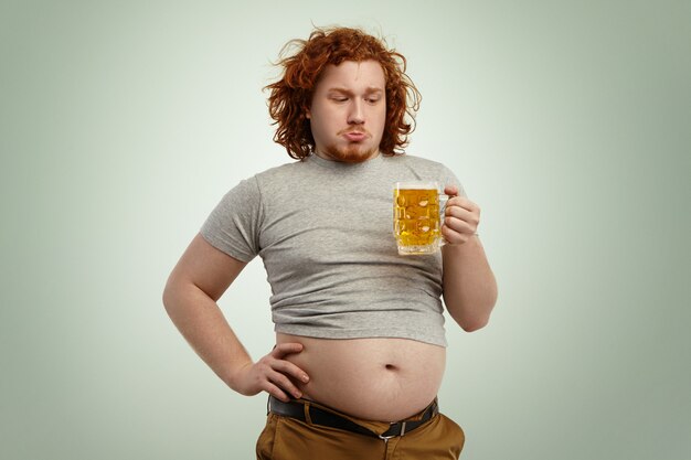 Personas, estilo de vida poco saludable, obesidad y glotonería. Sobrepeso gordo joven europeo con el pelo rojo rizado que sostiene un vaso de cerveza, sintiendo dudas al decidir si tomarlo o no después de una buena cena