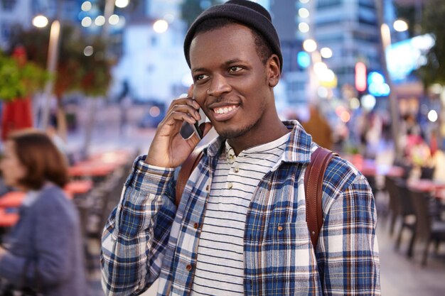 Personas, estilo de vida urbano, tecnología moderna y concepto de comunicación. Retrato al aire libre del hombre negro joven y guapo de moda que disfruta de un paseo nocturno por la ciudad, hablando por teléfono móvil con su amigo