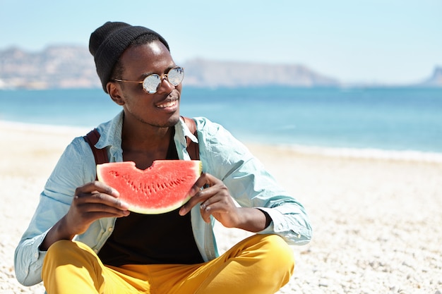 Personas y estilo de vida. Viaje y Turismo. Feliz relajado joven afroamericano hombre mochilero disfrutando de dulce sandía jugosa, sentado con las piernas cruzadas en Pebble Beach, sosteniendo fruta madura