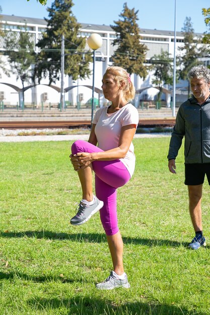 Personas maduras activas jubiladas en ropa deportiva haciendo ejercicio matutino en el césped del parque. Mujer en medias y zapatillas de deporte estirando las piernas. Concepto de jubilación o estilo de vida activo