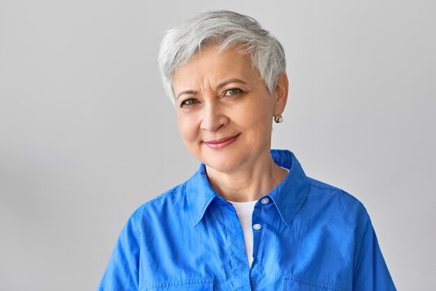 Personas maduras, concepto de envejecimiento y belleza. Alegre y atractiva pensionista de mediana edad con el pelo gris corto sonriendo, disfrutando de su jubilación, pasando el día en casa