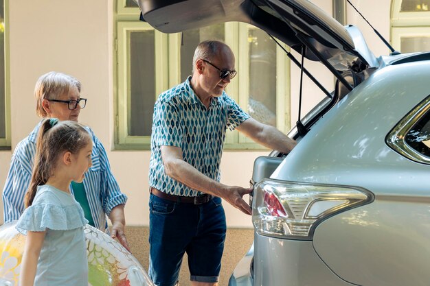 Personas mayores que se llevan a los niños de vacaciones en coche, viajando de vacaciones de verano en la playa. Abuelos y sobrina partiendo de aventura, poniendo bolsas de viaje e inflables en el maletero.