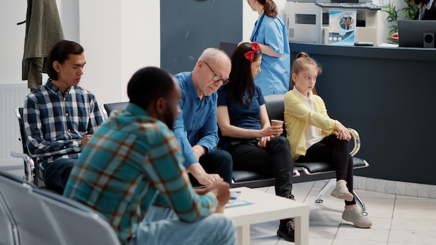 Personas multiétnicas esperando en el vestíbulo de recepción del hospital para asistir a una cita de control y hablar con el médico. Diversos pacientes sentados en la sala de espera en las instalaciones, apoyo de seguro médico.