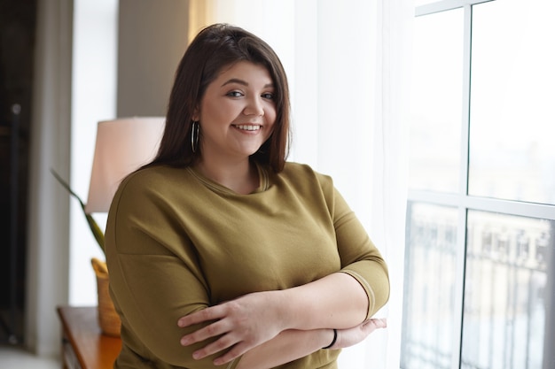 Personas, positividad corporal y concepto de estilo de vida. Imagen interior de adorable señorita gordita con sobrepeso relajándose en casa, de pie junto a la ventana, cruzando los brazos sobre su pecho, sonriendo ampliamente