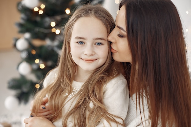 Personas que se preparan para la Navidad. Madre jugando con su hija. Familia sentada junto al árbol de Navidad. Niña con un vestido blanco.