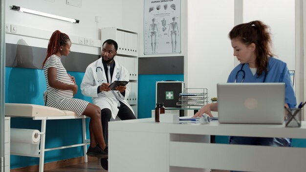 Personas que se reúnen en el consultorio médico para hablar sobre el embarazo y el parto, mujeres embarazadas que piden consejo a un obstetra. Médico conversando con paciente expectante y tomando notas.