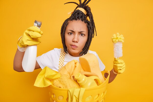 Personas quehaceres domésticos quehaceres domésticos y concepto de limpieza. Grave mujer afroamericana sostiene el cepillo y la botella de spray limpia el polvo vestido con uniforme casual aislado sobre fondo amarillo elimina la suciedad