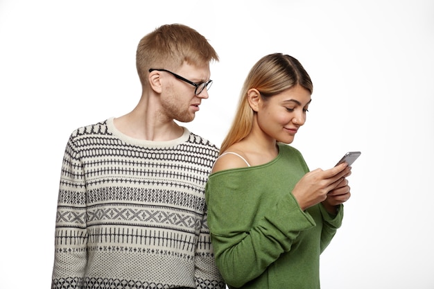 Personas, relaciones y tecnología moderna. Apuesto joven sin afeitar con gafas con mirada desconfiada mientras sospecha que su novia lo engaña, leyendo mensajes en su teléfono celular