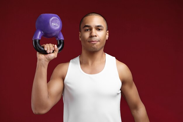 Personas, salud, deportes, fitness y powerlifting. Levantador de pesas afroamericano joven armado fuerte seguro levantando pesas 12 kg mientras hace ejercicio en el gimnasio, posando sobre fondo de pared de estudio rojo en blanco