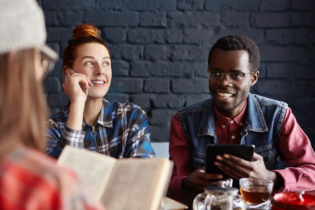 Personas, tecnología y comunicación. Grupo de tres jóvenes que tienen conversación en el café: mujer pelirroja hablando por teléfono celular, hombre africano con tableta electrónica