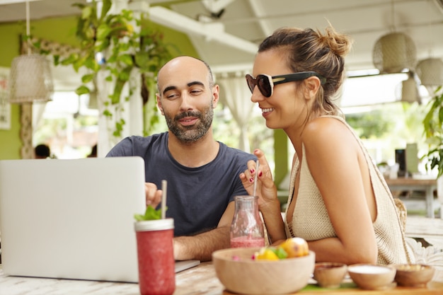 Personas, tecnología y comunicación. Pareja adulta con ordenador portátil en el café, sentado a la mesa con bebidas frescas. Hombre guapo mostrando algo a su novia en el cuaderno.