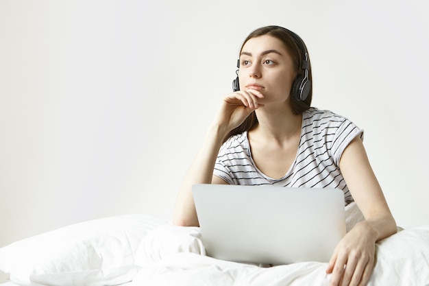 Personas, tecnología y concepto de estilo de vida moderno. Hermosa joven morena sentada en la ropa de cama blanca con la computadora portátil abierta en su regazo, usando auriculares mientras escucha un audiolibro en línea