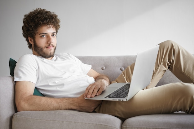 Personas, tecnología moderna y concepto de comunicación. Imagen de un chico guapo y elegante con barba sentado en el sofá con una computadora portátil en su regazo, disfrutando de una conexión inalámbrica a Internet de alta velocidad