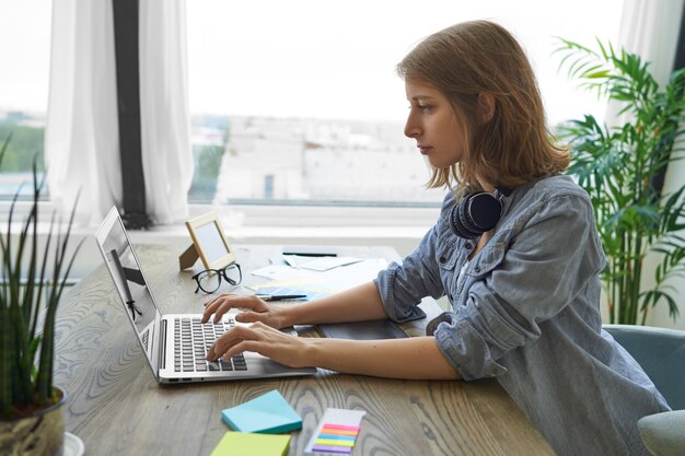 Personas, tecnología moderna y concepto de trabajo. Vista lateral del trabajador autónomo serio concentrado joven con auriculares alrededor de su cuello escribiendo en el teclado de la computadora portátil, sentado junto a la ventana en el escritorio de madera