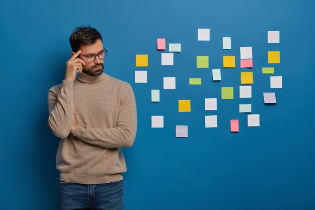 Personas, trabajo, concepto de pensamientos. Chico barbudo contemplativo mantiene el dedo en la sien, mira pensativamente a un lado, pone coloridas notas adhesivas en la pared