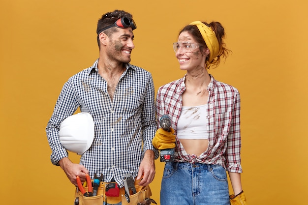 Personas, trabajo, ocupación y profesión. Dos talentosos jóvenes artesanos alegres que disfrutan trabajando juntos: chica guapa en gafas protectoras con taladro mirando a su guapo colega y sonriendo