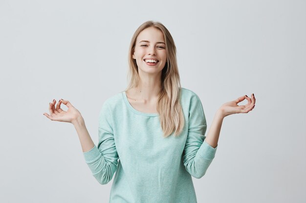 Personas, yoga y estilo de vida saludable. Joven rubia caucásica manteniendo los ojos cerrados mientras medita en el interior, practicando la tranquilidad, manteniendo los dedos en el gesto mudra, sonriendo ampliamente