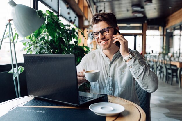 Perspectiva joven empresario en anteojos, sosteniendo una taza de café, hablando por teléfono