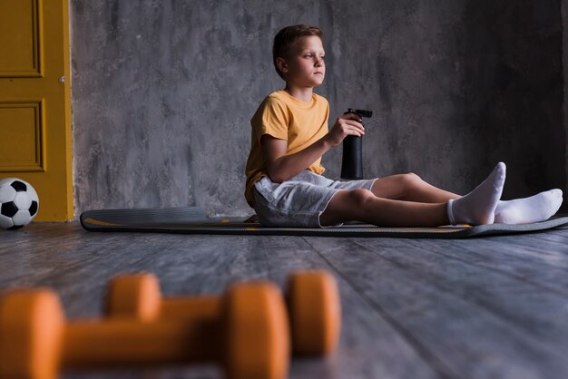 Pesas de gimnasia delante del muchacho que se sienta en la estera del ejercicio con la botella de agua