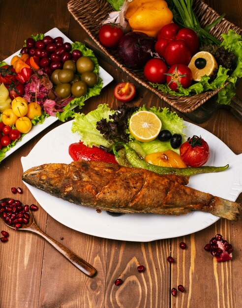 Pescado entero frito asado con verduras a la plancha y lechuga. En plato blanco decorado con turshu en mesa de madera.