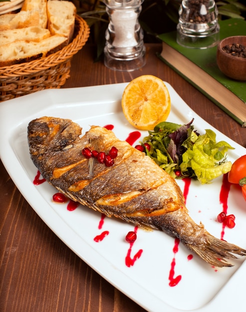 Pescado entero a la plancha con salsa amarilla, ensalada de verduras, limón y semillas de granada en un plato blanco