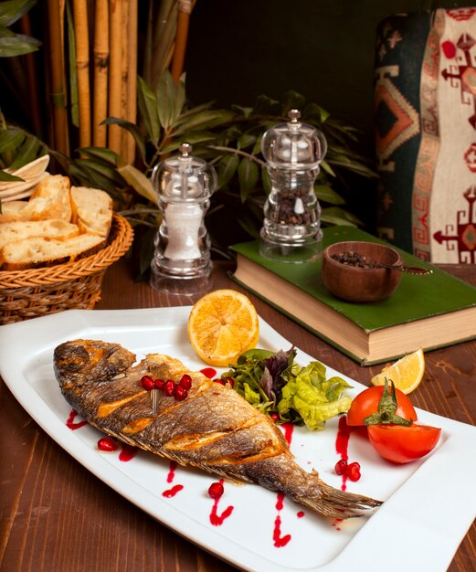 Pescado entero a la plancha con salsa amarilla, ensalada de verduras, limón y semillas de granada en un plato blanco