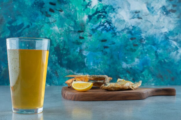 Pescado, rodajas de limón y un vaso de cerveza en una tabla, sobre la mesa de mármol.