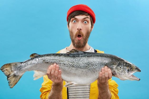 Pescador barbudo atónito vestido de manera informal sosteniendo un pez enorme que miraba con ojos saltones y la mandíbula caída se sorprendió al atrapar truchas o salmones tan grandes.